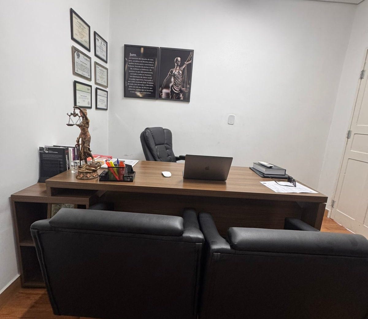 A sala de atendimento: mesa de madeira com laptop e a estátua da Justiça, poltrona preta atrás dela, duas poltronas de couro preto para quem é atendido e, na parede, os diplomas emoldurados e um quadro escuro.