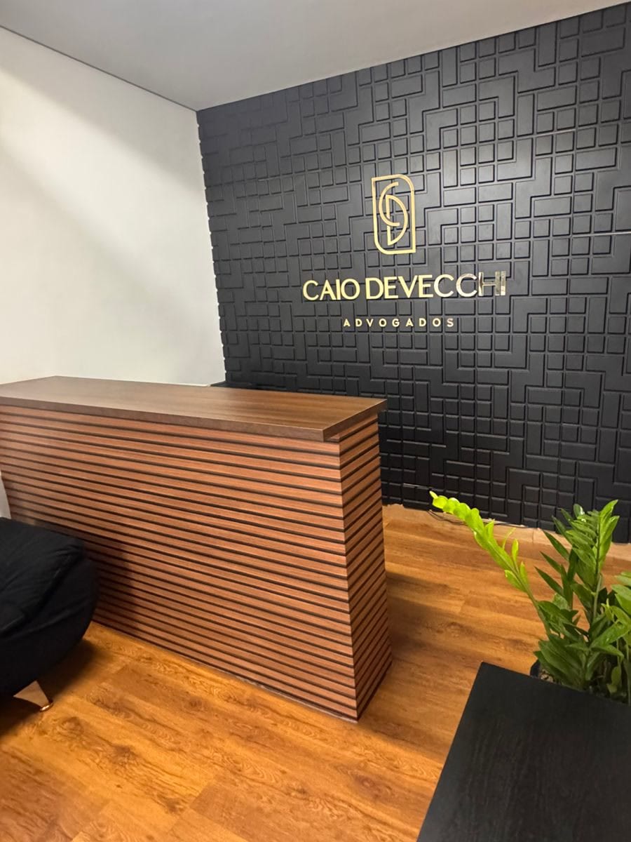 A recepção: painel preto em relevo com o monograma e o nome Caio Devecchi Advogados em dourado, o balcão ripado de madeira à esquerda e uma planta à direita, sobre o piso de madeira.