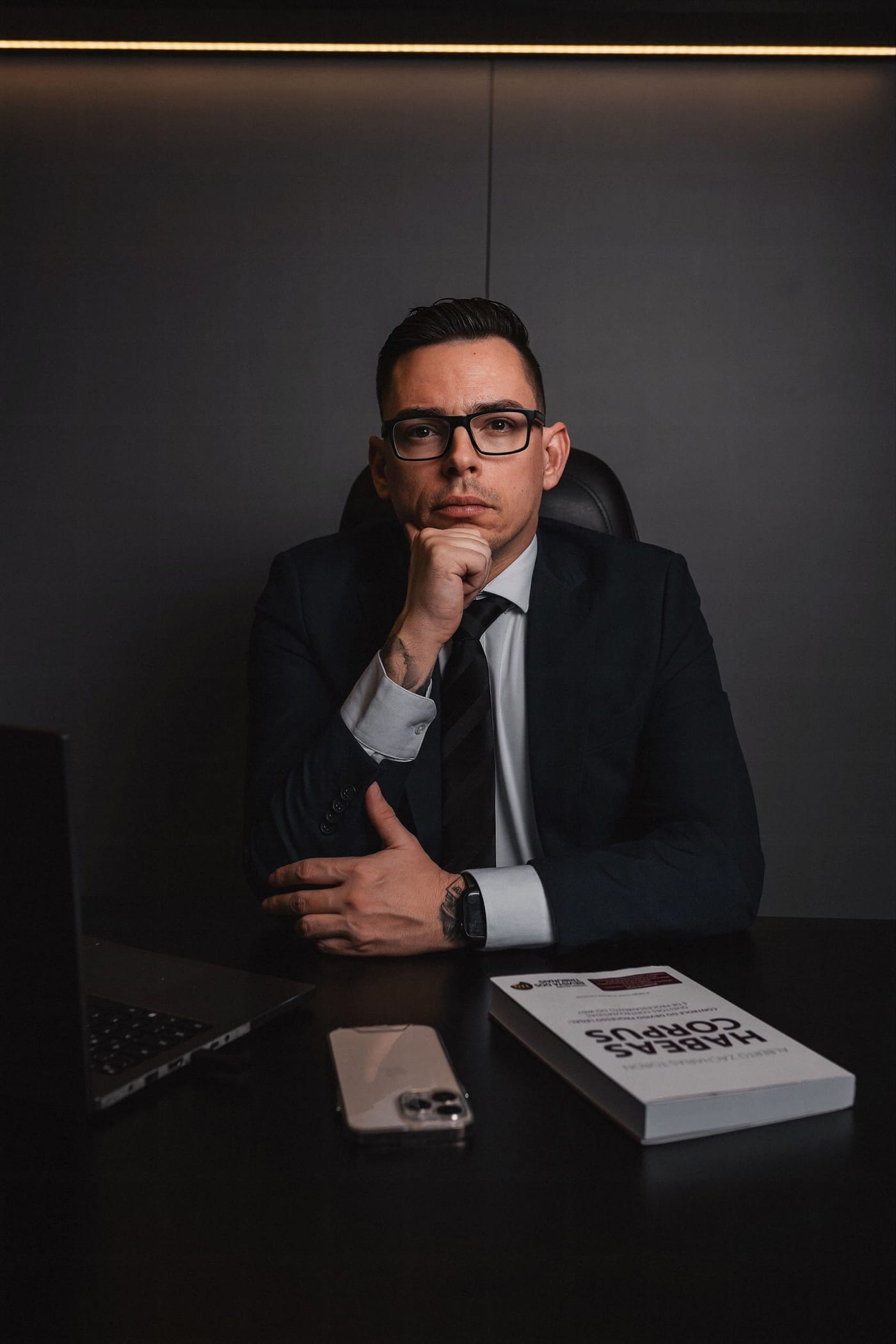 Caio Devecchi de frente, sentado à mesa, terno preto, gravata escura e óculos, a mão no queixo; sobre a mesa, um laptop, um celular e o livro Habeas Corpus.