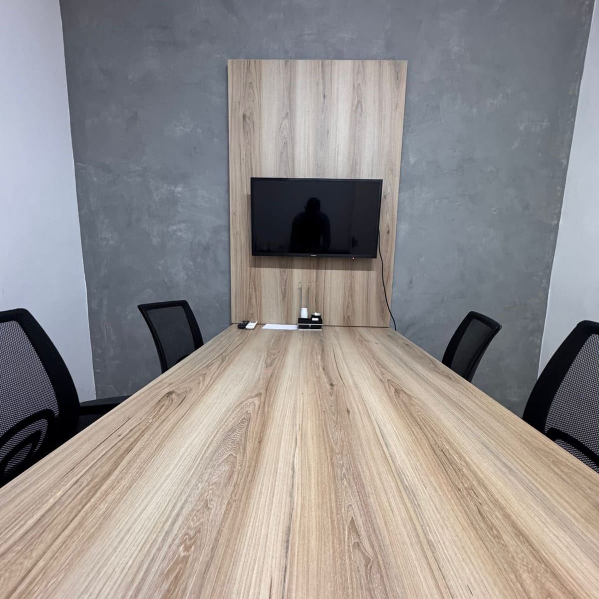 A sala de reuniões: mesa longa de madeira clara com cadeiras pretas e, na parede de cimento queimado, o painel de madeira com a TV.