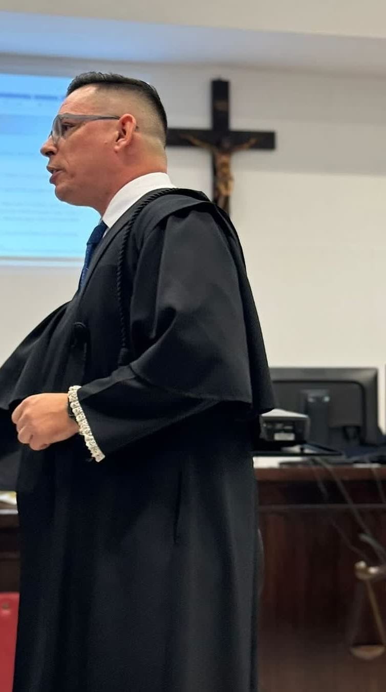 Caio Devecchi de toga preta com renda nos punhos, de pé e de perfil, falando no plenário do Tribunal do Júri; ao fundo, o crucifixo na parede e a tela do projetor.